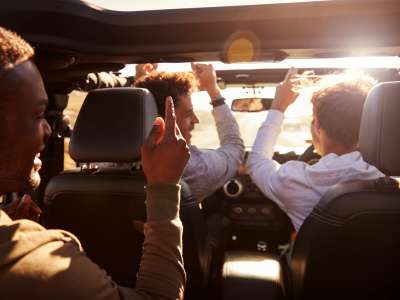 Drei Männer sitzen schick gekleidet in einem Auto und fahren bei bestem Wetter mit guter Laune durch die Gegend.