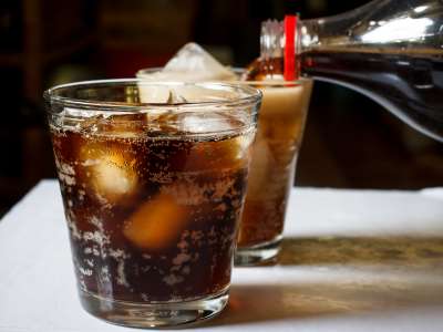 Zwei gläser Cola stehen auf einem Tisch und bei einem wird gerade nachgeschüttet. Beide sind mit Eiswürfel gefüllt.
