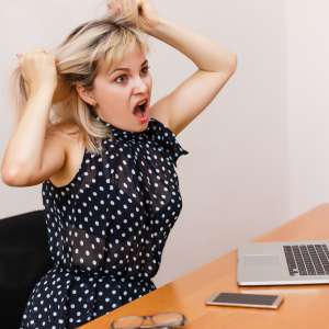 Frau in schwarzem Kleid, mit Weißen Punkten rauft sich die Haare. Sitzt dabei vor einem Tisch, auf dem Handy, Laptop und Brille liegen.