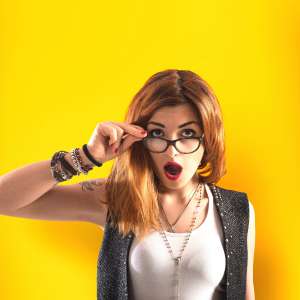 Vor einem Gelben Hintergrund steht eine Frau mit roten Haaren, Brille und weißem Oberteil.