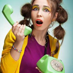 Eine knallig bunt gekleidete Frau steht mit ihrem Telefon in der Hand da und schaut sehr überrascht.
