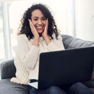 Eine Frau sitzt auf der Couch mit ihrem Laptop in der Hand und schaut sehr Erstaunt auf den Laptop.