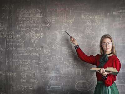 Eine Junge Lehrerin steht vor einer Tafel mit vielen verschiedenen Formeln darauf.