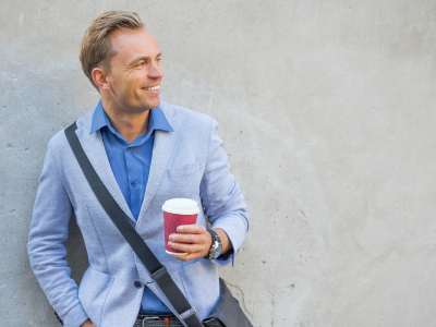 Attraktiver Mann im blauen Anzug hält eine Kaffeetasse und lächelt, während er sich an eine Betonwand lehnt - Tipps zum Mann verführen.