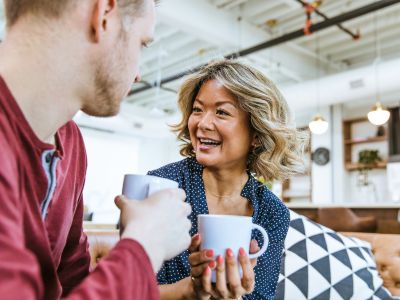 Eine blonde Frau mit einer Kaffeetasse sitz einem Mann gegenüber der auch eine Kaffeetasse hält. Beide lernen sich kennen