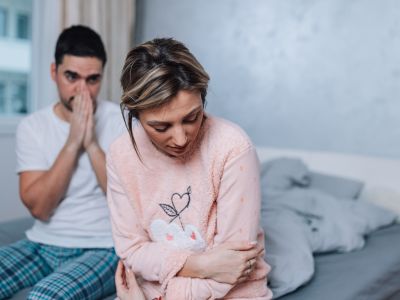 Eine Frau sitzt besorgt am Bettrand, die Arme verschränkt, während ein Mann hinter ihr sitzt und sich schockiert die Hand vor den Mund hält. Die Szene spiegelt die Bindungsangst in der Kennenlernphase eines Paares wider.