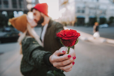 Rote Rose in der Hand einer Frau, Paar küsst sich im Hintergrund – Symbol für Liebe und Leidenschaft