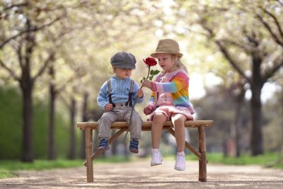 Zwei stilvoll gekleidete Kinder auf einer Bank im blühenden Park, das Mädchen überreicht dem Jungen eine rote Rose – Symbol für romantische Geste und echte Zuneigung.