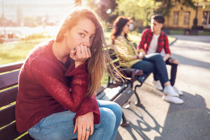 Eine junge Frau sitzt eifersüchtig und nachdenklich allein auf einer Parkbank, während ihr Freund im unscharfen Hintergrund mit seiner Ex-Partnerin sitzt, was die Zwickmühle einer neuen Partnerin symbolisiert.