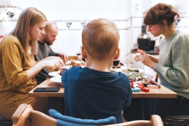 Ein Kind sitzt im Fokus am Esstisch, während der Vater mit seiner Ex-Partnerin und der neuen Freundin isst, was die Herausforderung darstellt, wenn der Mann mit der Ex wohnt.