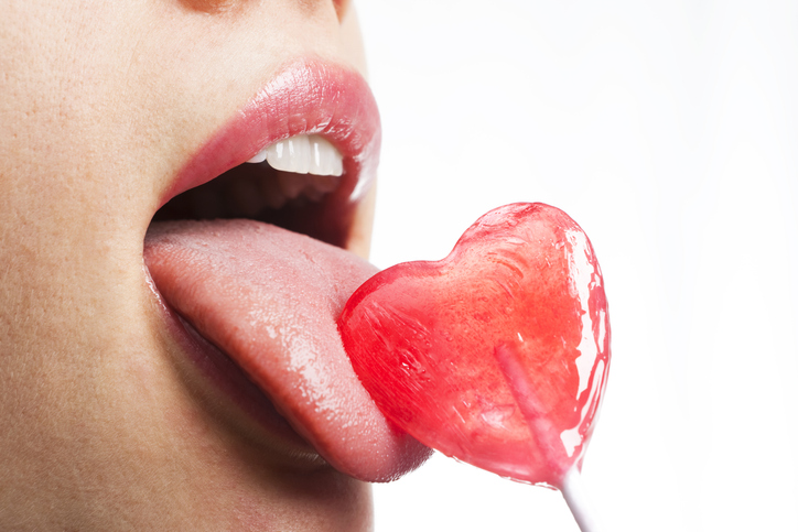 Nahaufnahme eines Mundes mit glänzenden Lippen, der einen roten herzförmigen Lutscher leckt.