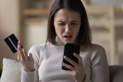 Eine Frau hält frustriert eine Kreditkarte und ihr Handy in den Händen, weil sie für die Nutzung einer Dating-App zahlen muss.