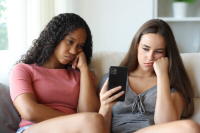 Zwei Freundinnen sitzen genervt auf dem Sofa und schauen auf ein Smartphone.