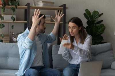 Paar streitet auf dem Sofa über eine Rechnung; erhobene Hände, angespannte Stimmung – verpasster Zeitpunkt fürs Geldgespräch bei Gehaltsunterschieden in der Beziehung.