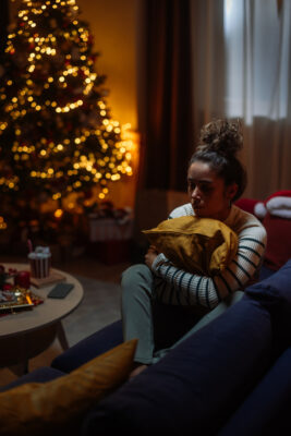 Frau sitzt traurig auf dem Sofa neben leuchtendem Weihnachtsbaum – Weihnachten allein, Weihnachtsblues und Einsamkeit.