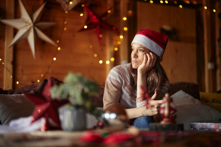 Nachdenkliche Frau mit Weihnachtsmütze sitzt allein im festlich geschmückten Wohnzimmer – Single an Weihnachten, Weihnachtsblues.