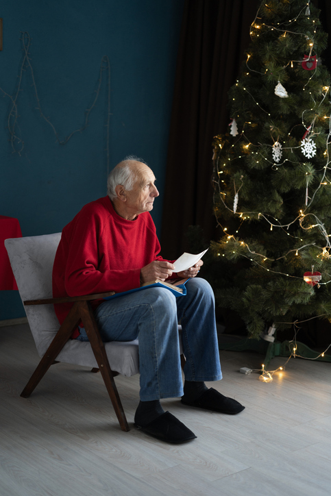 Älterer Mann sitzt allein neben dem Weihnachtsbaum und liest einen Brief – Einsamkeit an Weihnachten, mentale Gesundheit.