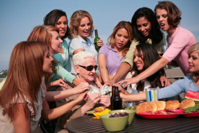 Älterer Mann mit vielen jungen Frauen am Tisch, hält Geldscheine – Symbolbild für Sugar Daddy und Sugar Babies