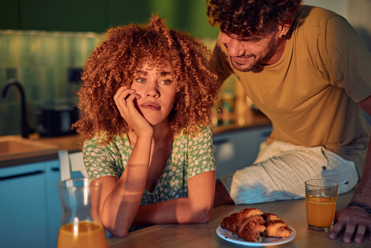 Frau wirkt nachdenklich und genervt am Tisch, Mann versucht zu reden – unrealistische Erwartungen beim Dating und „Einhorn“-Suche