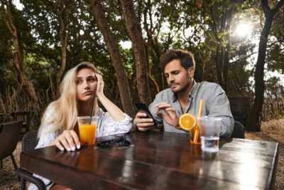 Eine gelangweilte Frau blickt auf ein Smartphone nutzenden Mann, während sie an einem Tisch sitzen.
