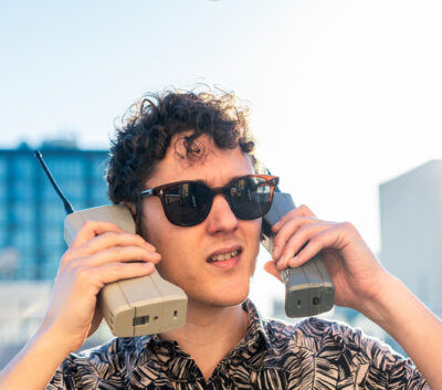 Ein Mann mit Sonnenbrille und Hemd hält zwei alte, klobige Mobiltelefone an die Ohren, mit einem verwirrten Ausdruck.