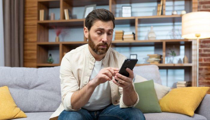 Nachdenklicher Mann mit Dating-Burnout schaut auf sein Smartphone Ein nachdenklicher Mann sitzt auf einem Sofa und schaut auf sein Smartphone, während er eine Dating-App benutzt.